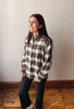 A woman modeling a long-sleeve plaid button-up shirt in brown, beige, and white, paired with black pants, standing in a room with a light gray wall and wooden floor.