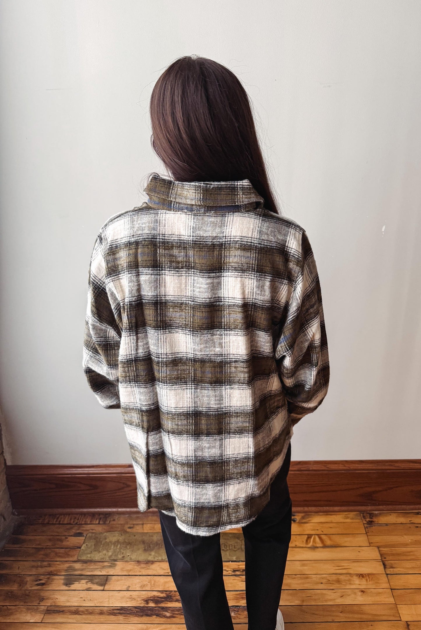 A person viewed from behind wearing an oversized plaid button-up shirt in brown, beige, and white, paired with black pants, standing on a wooden floor against a plain wall.