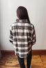 A person viewed from behind wearing an oversized plaid button-up shirt in brown, beige, and white, paired with black pants, standing on a wooden floor against a plain wall.