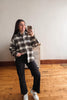 A woman takes a mirror selfie wearing a black, white, and gray plaid button-down shirt, black wide-leg pants, and white sneakers in a room with a wooden floor and white wall.