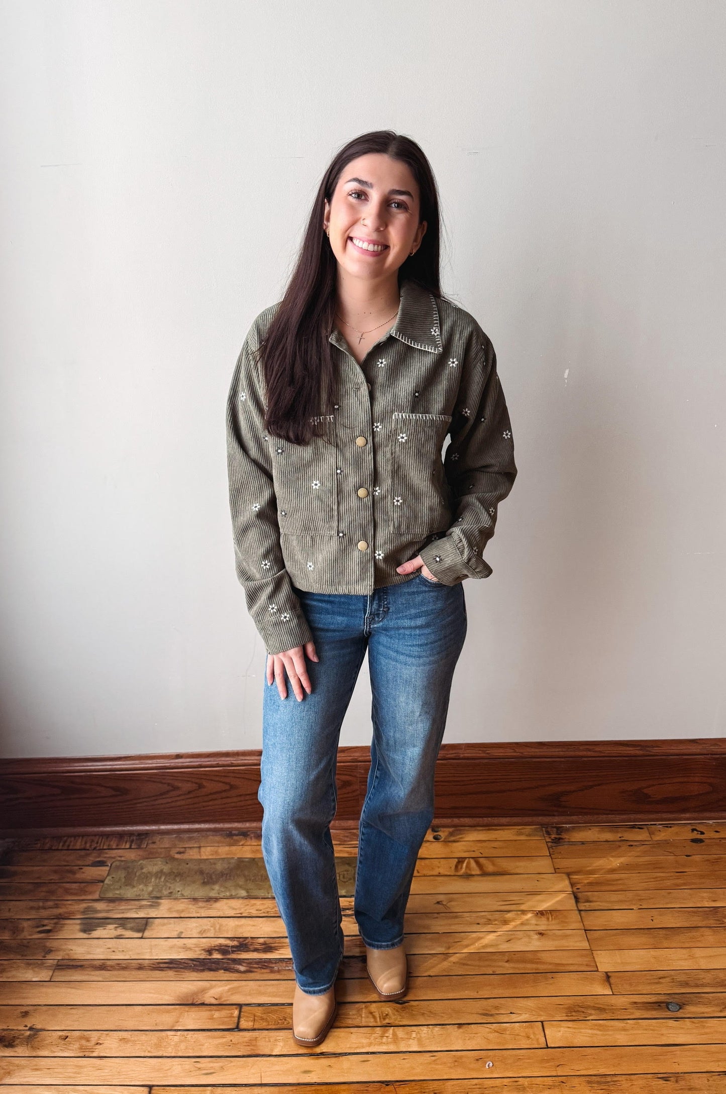A woman stands in a room with a light gray wall and wooden floor, wearing a muted olive green western-style button-up shirt with silver embroidery, blue jeans, and tan boots.