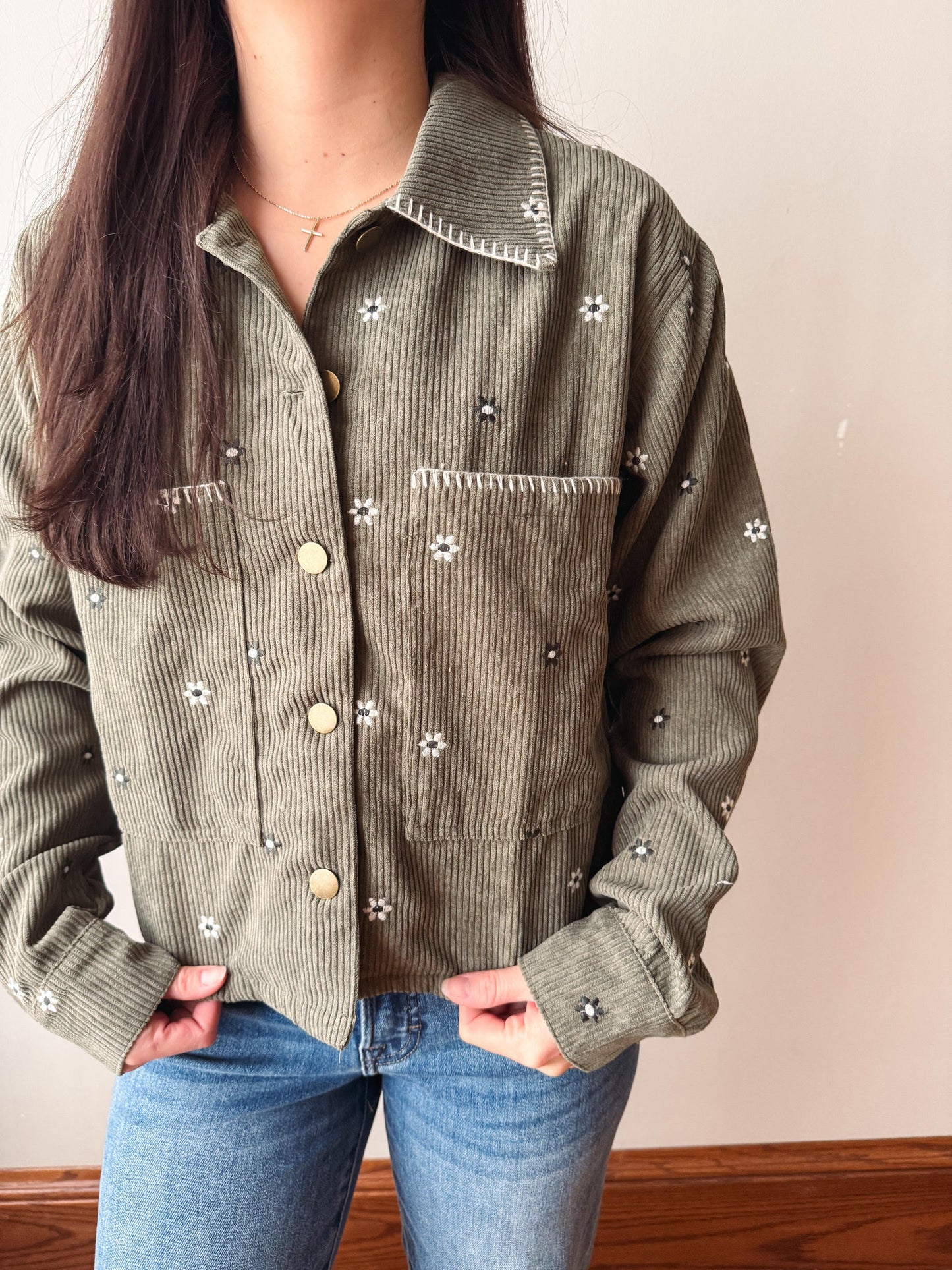 A person wearing an olive green corduroy button-up shirt jacket with embroidered white flowers and blue jeans, standing against a plain beige wall.