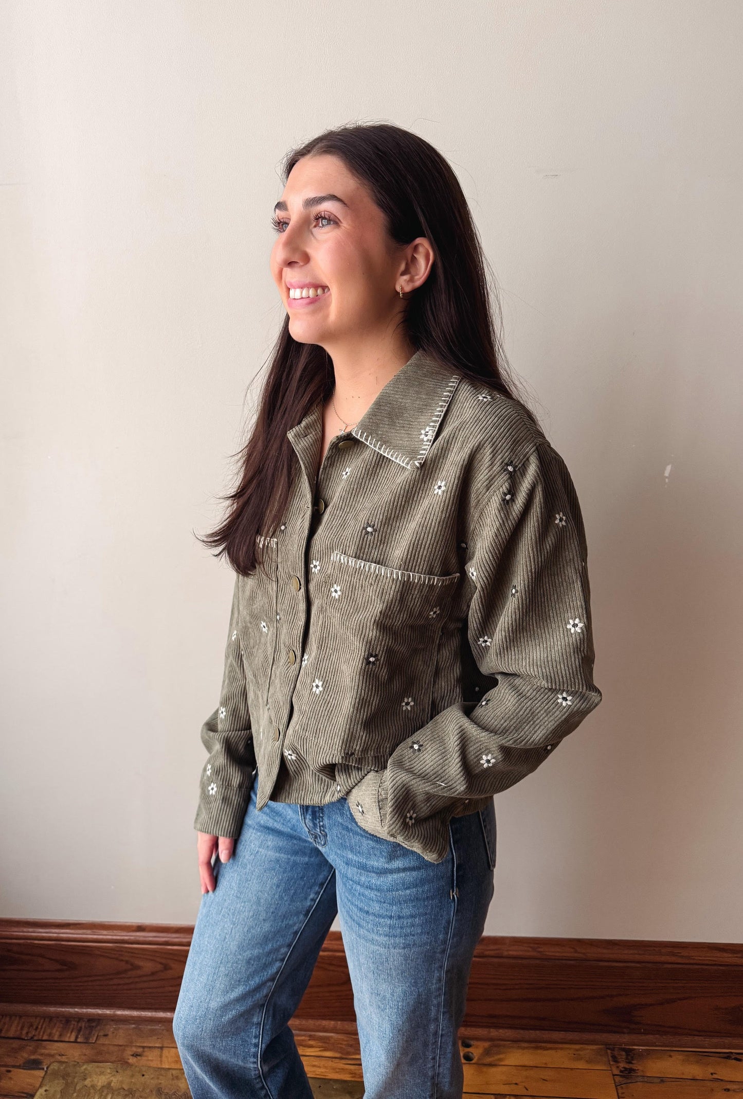 A woman wearing a sage green corduroy button-up shirt with white embroidered details, paired with blue jeans, standing against a plain beige wall.