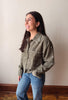 A woman wearing a sage green corduroy button-up shirt with white embroidered details, paired with blue jeans, standing against a plain beige wall.
