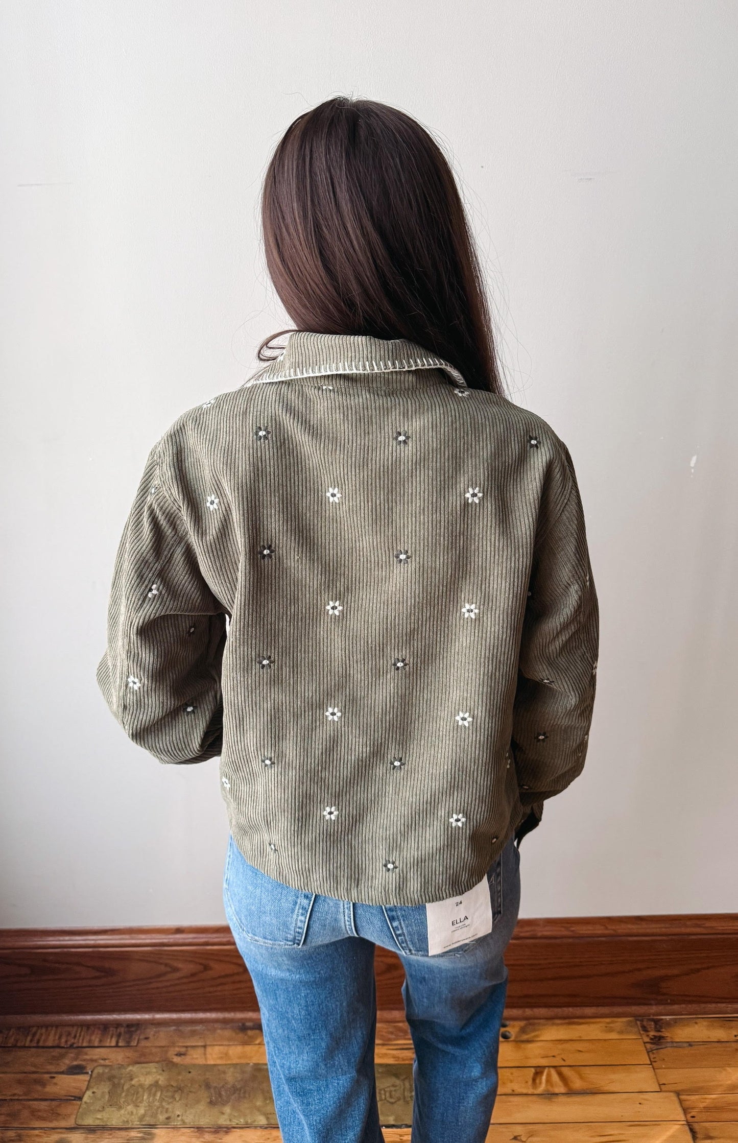 A person viewed from behind wearing a corduroy jacket in a muted olive green color with small white embroidered floral details, paired with blue jeans, standing against a plain off-white wall.