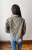 A person viewed from behind wearing a corduroy jacket in a muted olive green color with small white embroidered floral details, paired with blue jeans, standing against a plain off-white wall.