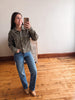 A woman takes a mirror selfie in a room with hardwood floors and a light-colored wall, wearing a brown western-style denim jacket with silver embroidery, blue jeans, and tan boots.