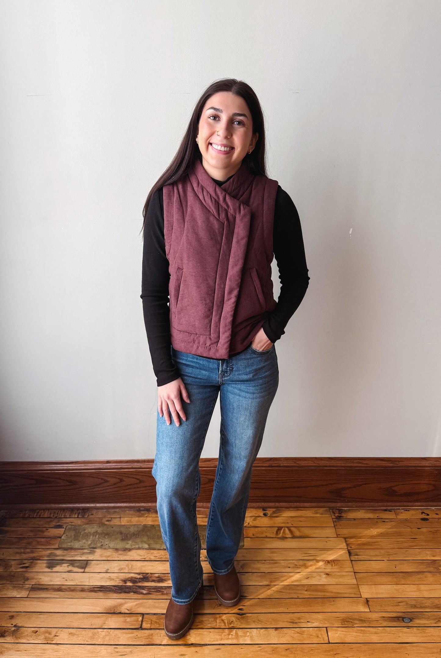 A woman modeling a sleeveless, quilted dusty rose vest over a black long-sleeve shirt and blue jeans, standing on a wooden floor against a plain off-white wall.