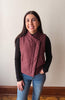 A woman wearing a dusty rose wrap-style quilted vest over a black long-sleeve shirt and blue jeans, standing against a plain off-white wall.