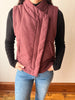 A person wearing a sleeveless dusty rose corduroy vest over a black long-sleeve shirt and blue jeans, standing indoors against a plain wall.
