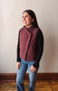 A woman wearing a sleeveless, quilted wrap-style vest in a dusty burgundy color over a black long-sleeve shirt and blue jeans, standing against a plain beige wall.