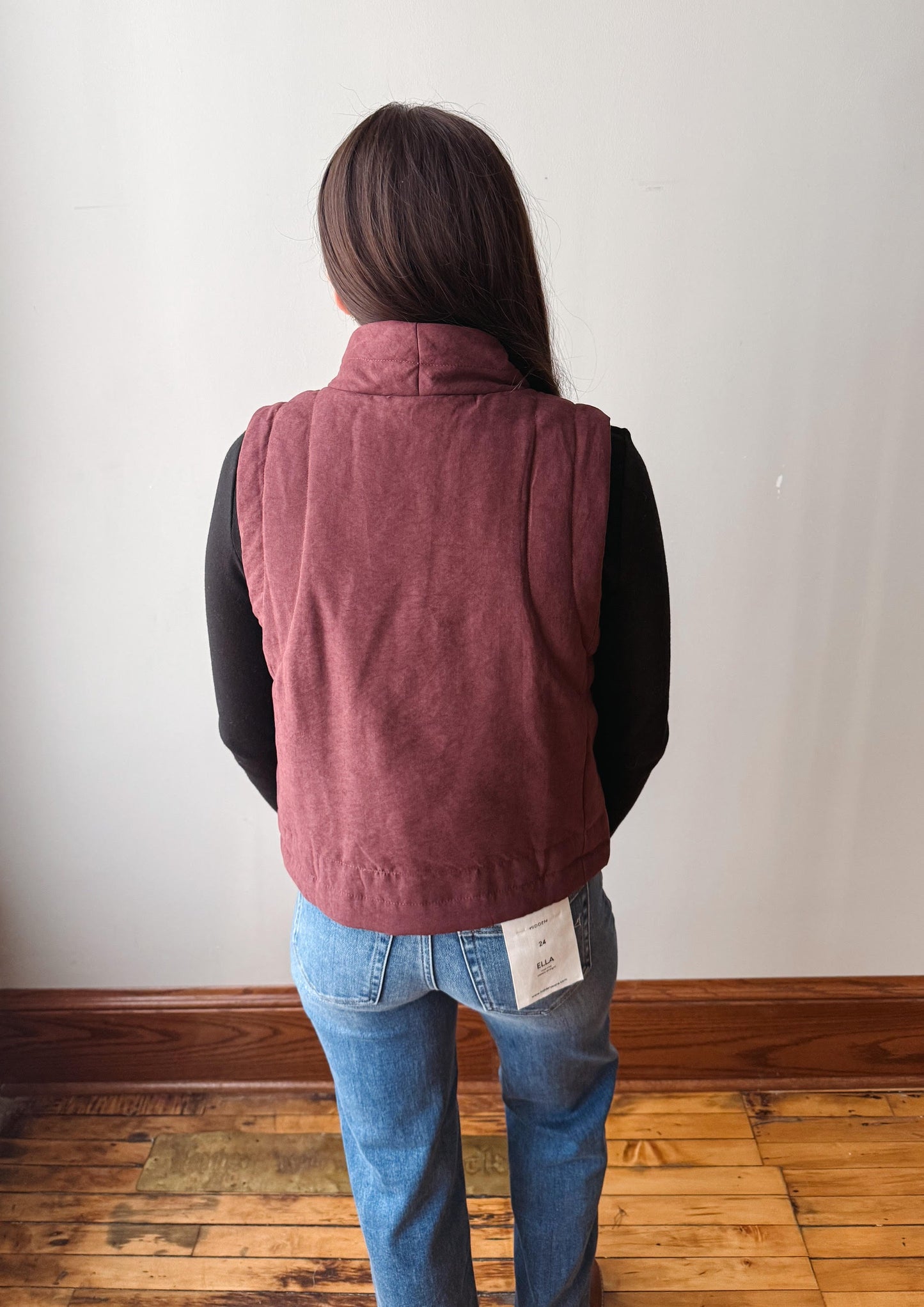 A person viewed from behind wearing a dusty rose corduroy vest over a black long-sleeve shirt and blue jeans, standing on a wooden floor against a plain white wall.