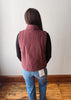 A person viewed from behind wearing a dusty rose corduroy vest over a black long-sleeve shirt and blue jeans, standing on a wooden floor against a plain white wall.
