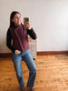 A woman taking a mirror selfie in an indoor room with hardwood floors and a white wall, wearing a sleeveless quilted burgundy vest over a black long-sleeve shirt and blue jeans.