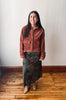 A woman stands in a room with a white wall and wooden floor, wearing a rust-brown cropped corduroy jacket over a dark green and brown patterned maxi skirt with tan ankle boots.