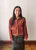 A woman modeling a rust-brown corduroy button-up shirt jacket paired with a green floral skirt, standing against a plain off-white wall with wooden flooring visible.