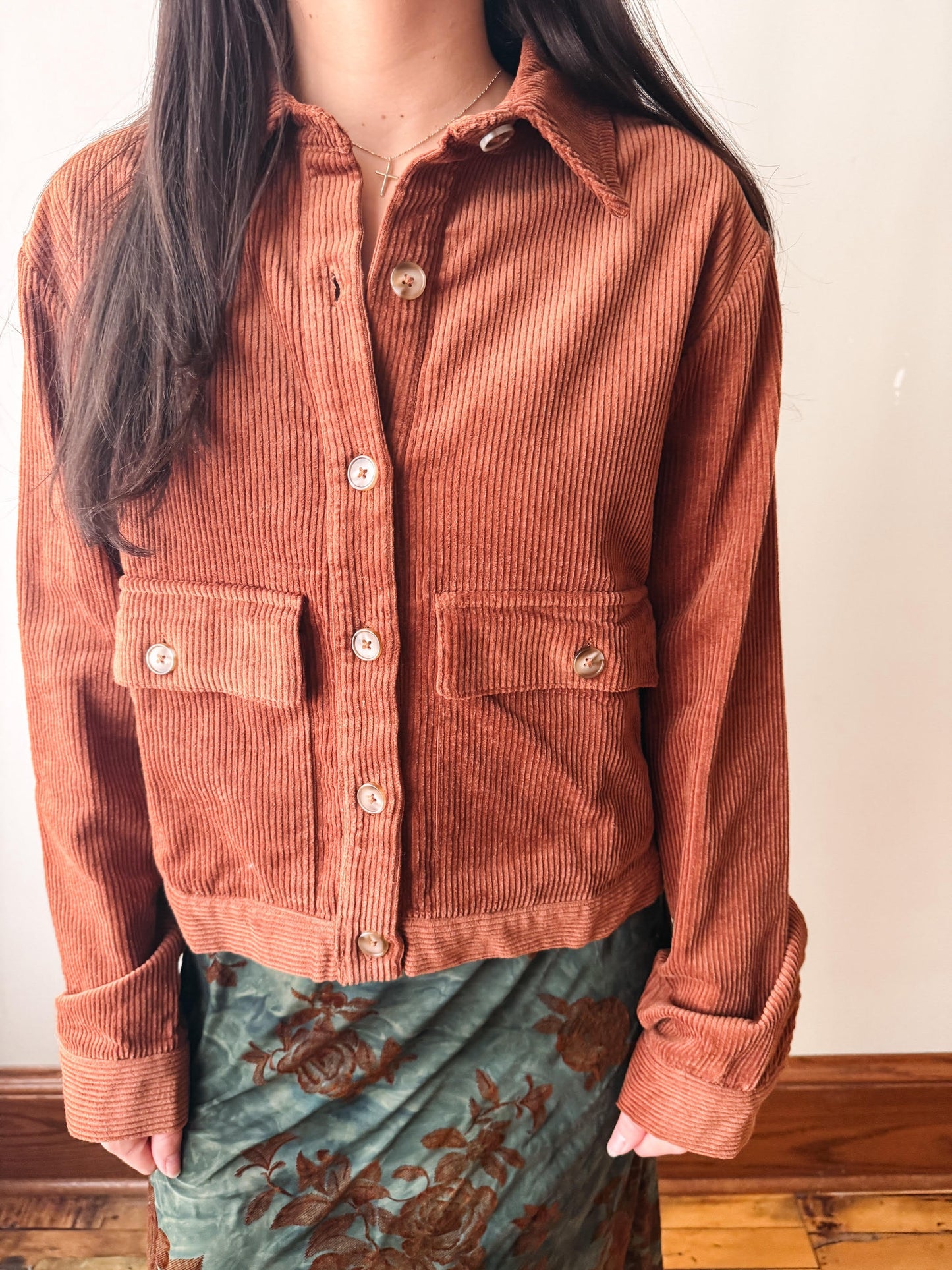 A person wearing a cropped corduroy button-front jacket in a muted terracotta/rust color, paired with a green floral skirt, standing indoors against a plain light wall.