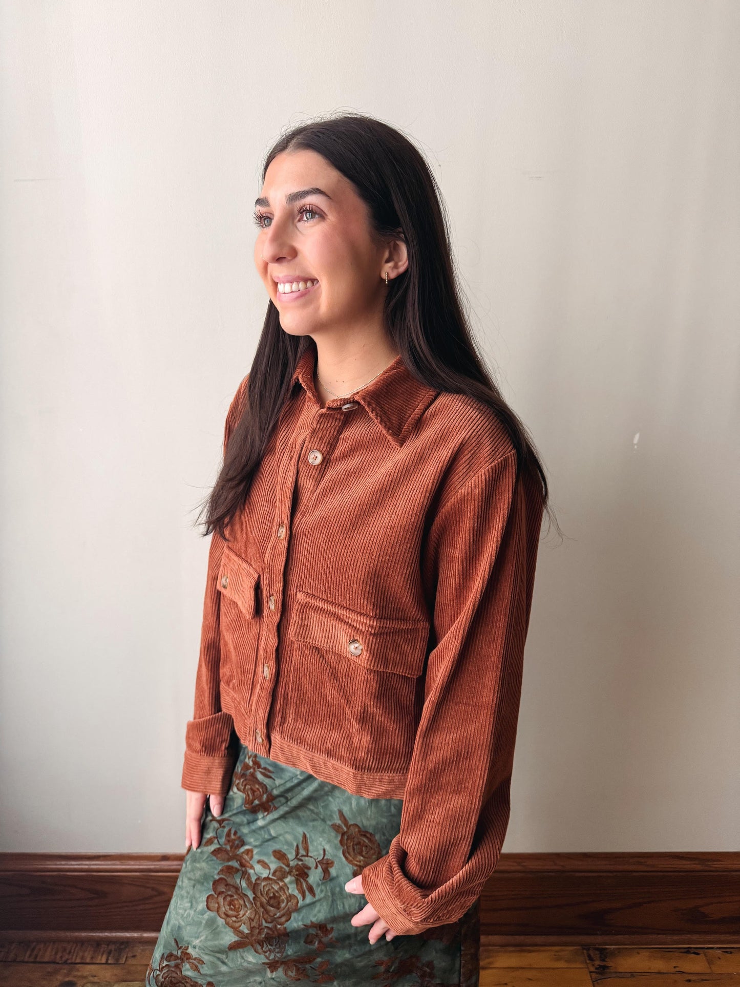 A woman wearing a rust-brown corduroy button-up shirt jacket and a green floral skirt, standing against a plain off-white wall.