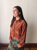 A woman wearing a rust-brown corduroy button-up shirt jacket and a green floral skirt, standing against a plain off-white wall.