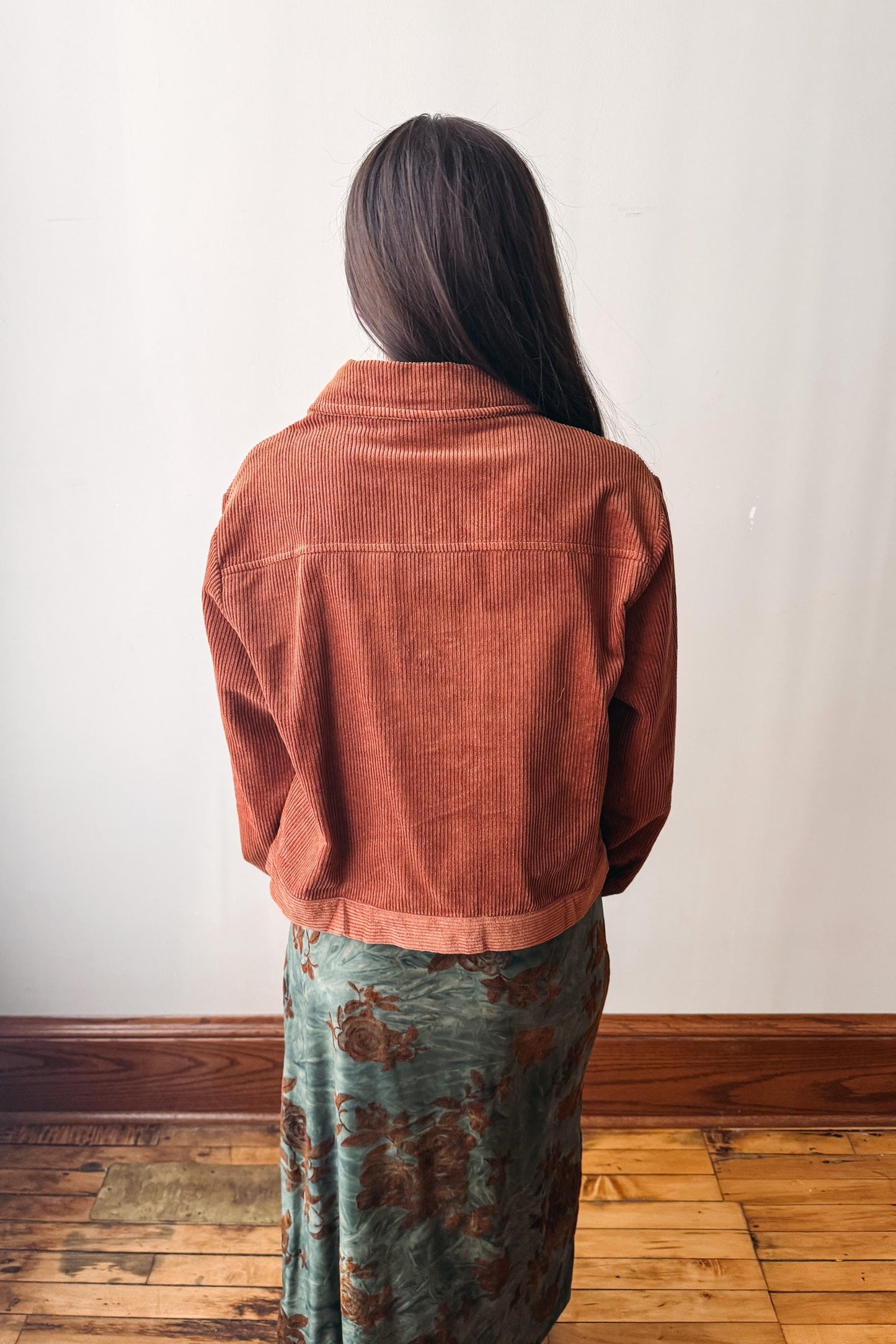 A person viewed from behind wearing a rust-orange corduroy cropped jacket over a green floral patterned skirt, standing on a wooden floor against a plain white wall.