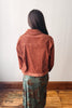 A person viewed from behind wearing a rust-orange corduroy cropped jacket over a green floral patterned skirt, standing on a wooden floor against a plain white wall.