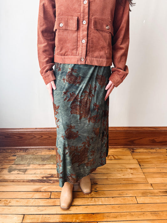 A person wearing a rust-colored corduroy jacket and a green floral maxi skirt, standing on a wooden floor against a white wall.