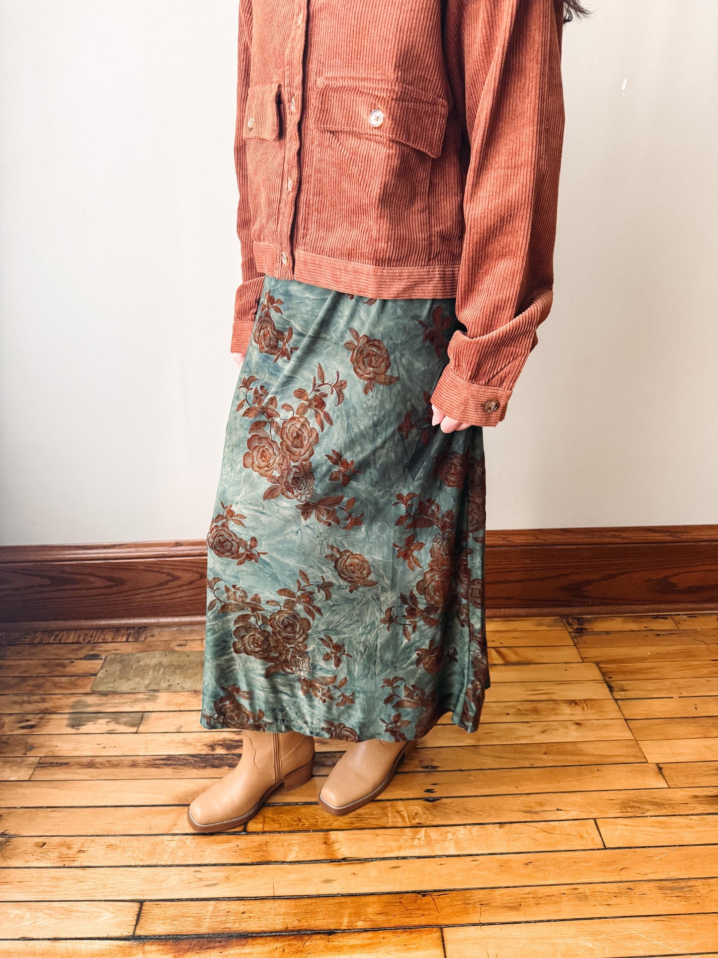 A person wearing a rust-colored corduroy jacket and a teal-green midi skirt with a brown floral pattern, standing on a wooden floor against a white wall.