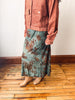 A person wearing a rust-colored corduroy jacket and a teal-green midi skirt with a brown floral pattern, standing on a wooden floor against a white wall.