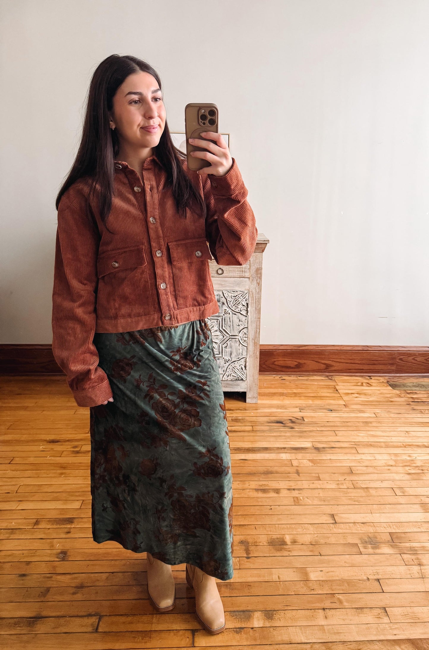A woman takes a mirror selfie wearing a rust-brown cropped suede jacket and a dark green patterned midi skirt, standing in a room with light wood floors and a white wall.