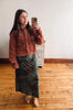 A woman takes a mirror selfie wearing a rust-brown cropped suede jacket and a dark green patterned midi skirt, standing in a room with light wood floors and a white wall.