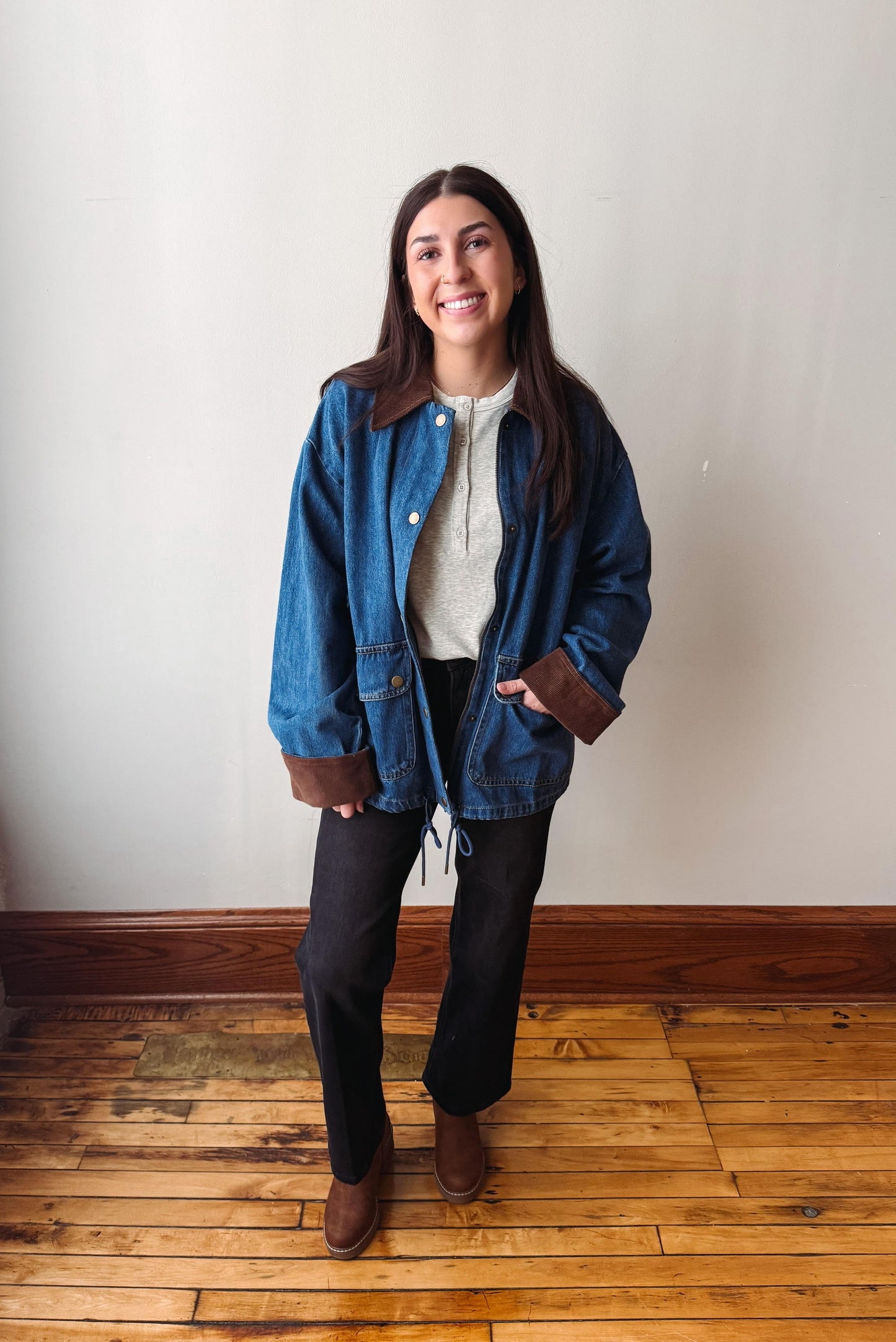 A woman stands in a room with a beige wall and wooden floor, wearing a blue denim jacket with brown suede-like trim over a cream top and black pants.