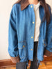 A person wearing a blue denim jacket with brown corduroy collar and cuffs, layered over a light gray shirt and dark pants.