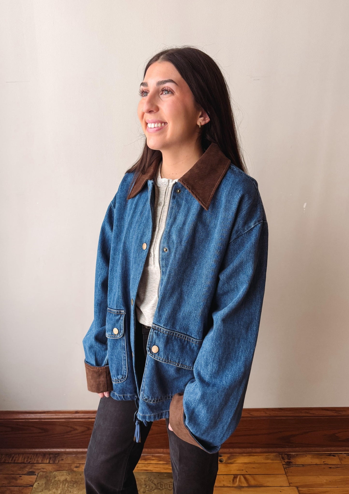 A woman wearing a blue denim jacket with brown suede-like collar and cuffs, layered over a white top and dark pants, standing against a plain beige wall with wooden flooring.