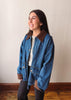 A woman wearing a blue denim jacket with brown suede-like collar and cuffs, layered over a white top and dark pants, standing against a plain beige wall with wooden flooring.