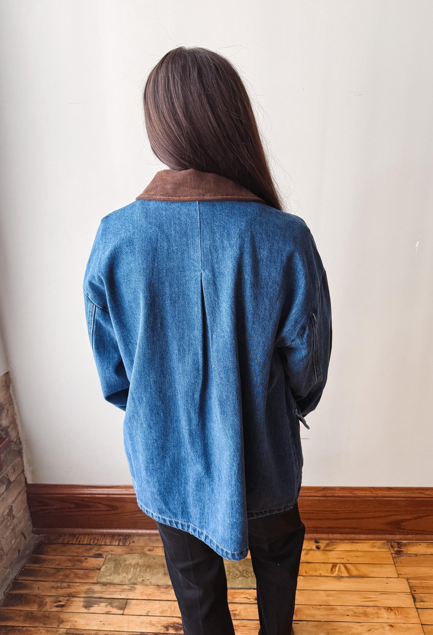 A person viewed from behind wearing a blue denim jacket with a brown collar, standing on a wooden floor against a plain off-white wall.