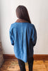 A person viewed from behind wearing a blue denim jacket with a brown collar, standing on a wooden floor against a plain off-white wall.