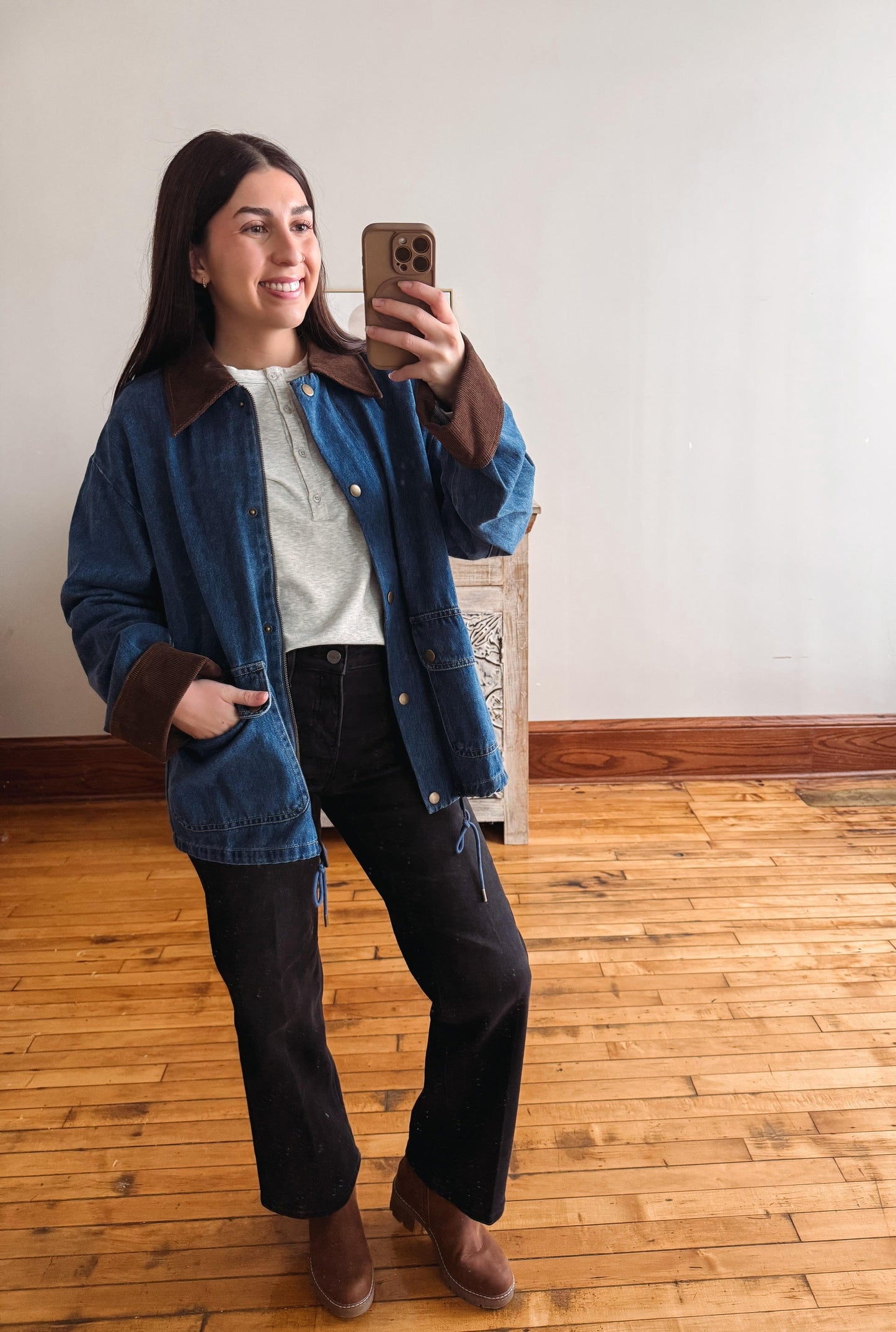 A woman taking a mirror selfie in an indoor room with wooden floors and a plain wall, wearing a blue denim jacket with brown suede-like collar and cuffs, a white top, black pants, and brown ankle boots.