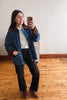 A woman taking a mirror selfie in an indoor room with wooden floors and a plain wall, wearing a blue denim jacket with brown suede-like collar and cuffs, a white top, black pants, and brown ankle boots.