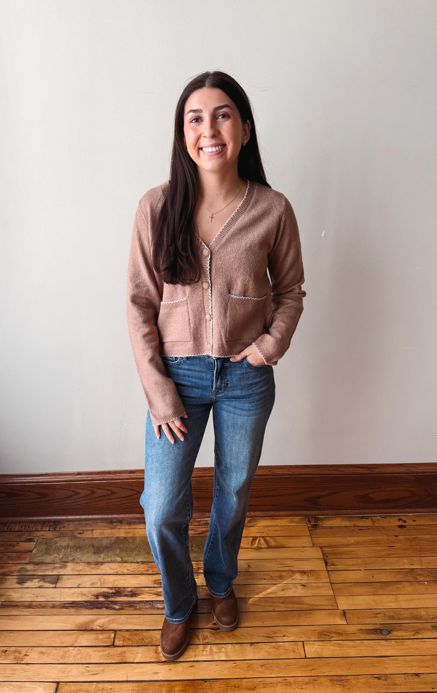 A woman stands in a room with a light gray wall and wooden floor, wearing a taupe-brown cropped cardigan, blue jeans, and brown shoes.