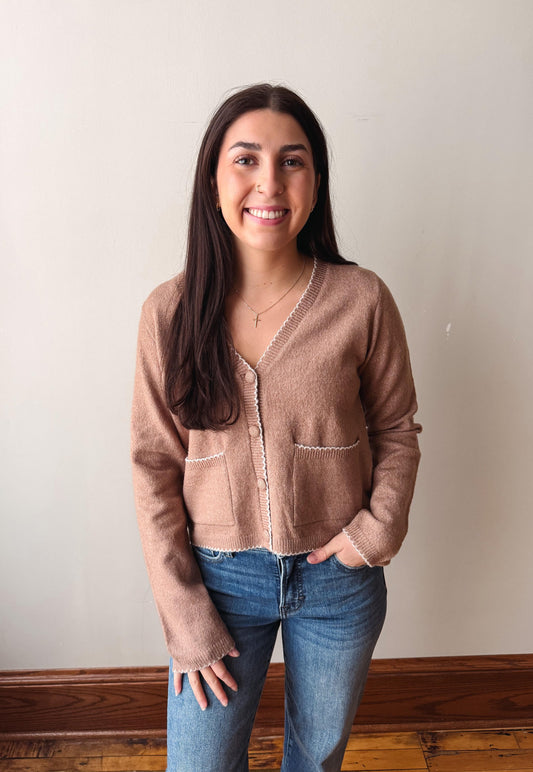 A woman with long dark hair stands indoors wearing a cropped taupe-brown knit cardigan with white contrast stitching and blue jeans, against a plain off-white wall with a wooden baseboard.