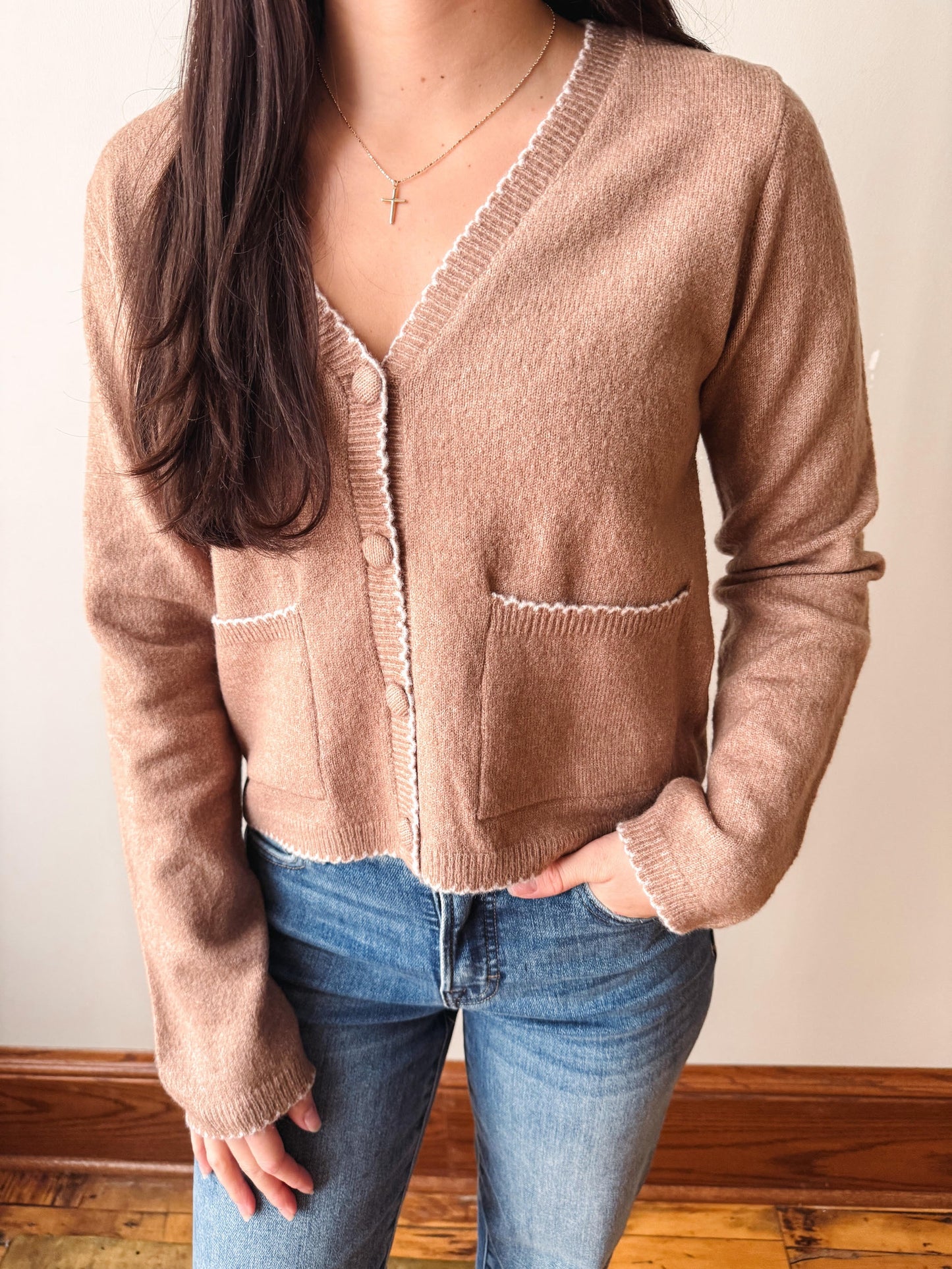 A woman wearing a cropped, long-sleeve knit cardigan in a warm taupe-brown color with white contrast stitching and button details, paired with blue jeans.