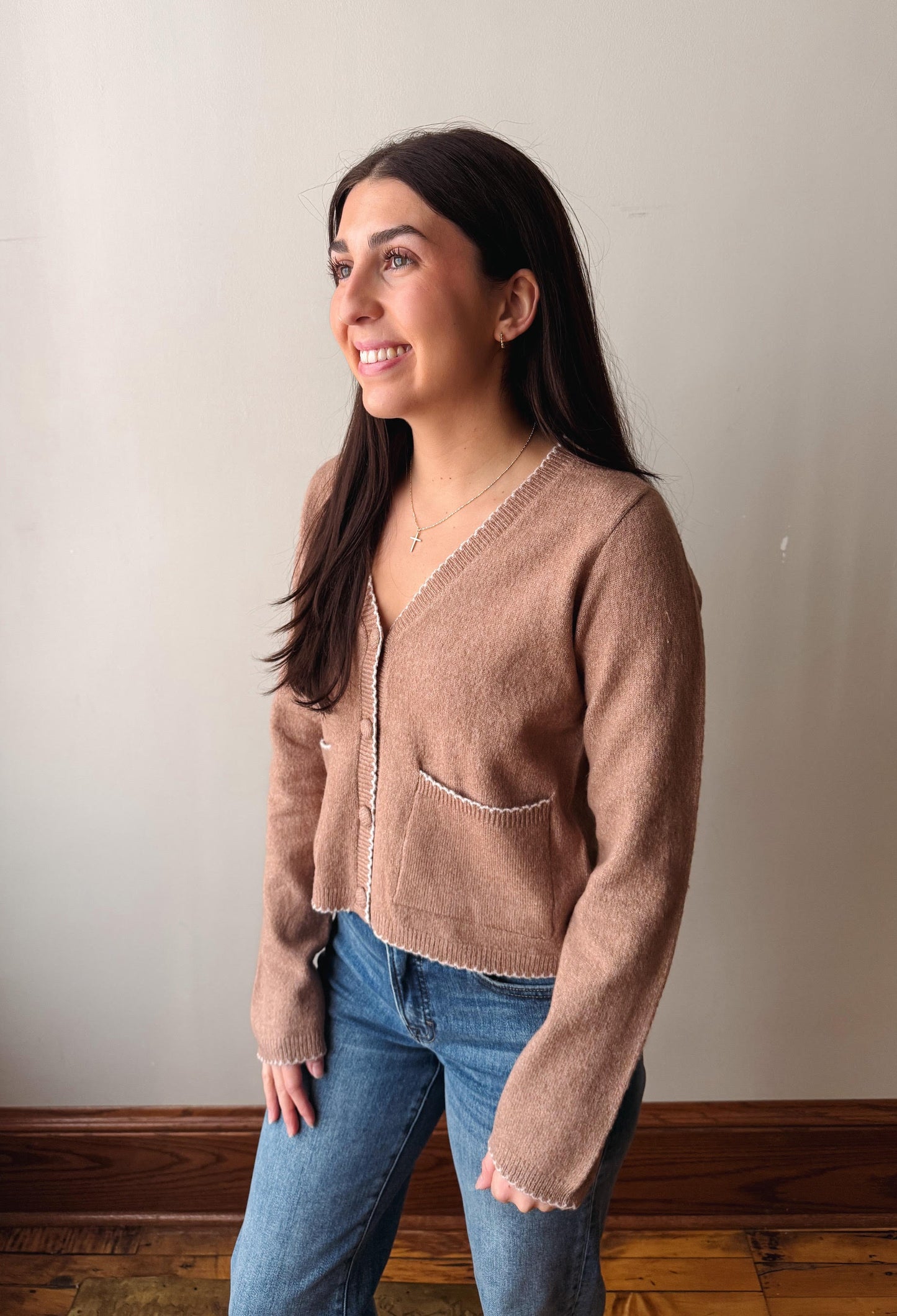 A woman wearing a light brown fuzzy knit cardigan with frayed edges and blue jeans, standing against a plain light gray wall.