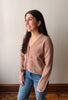 A woman wearing a light brown fuzzy knit cardigan with frayed edges and blue jeans, standing against a plain light gray wall.