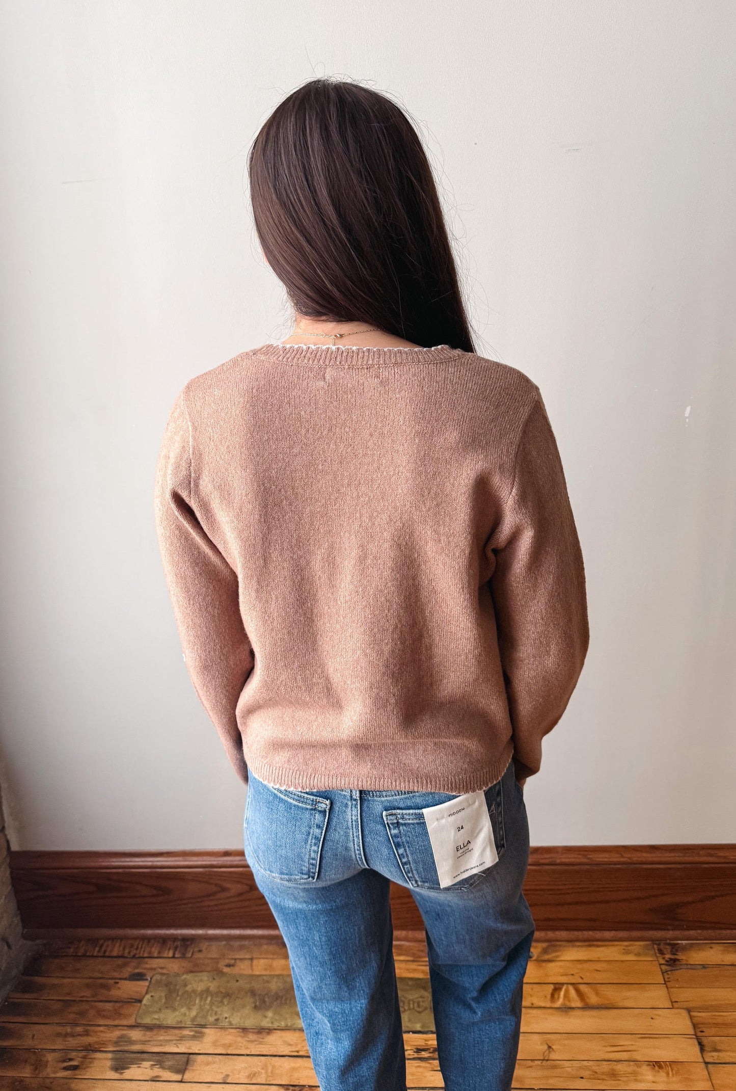 A woman viewed from behind wearing a dusty rose knit sweater and blue jeans, standing on a wooden floor against a plain off-white wall.