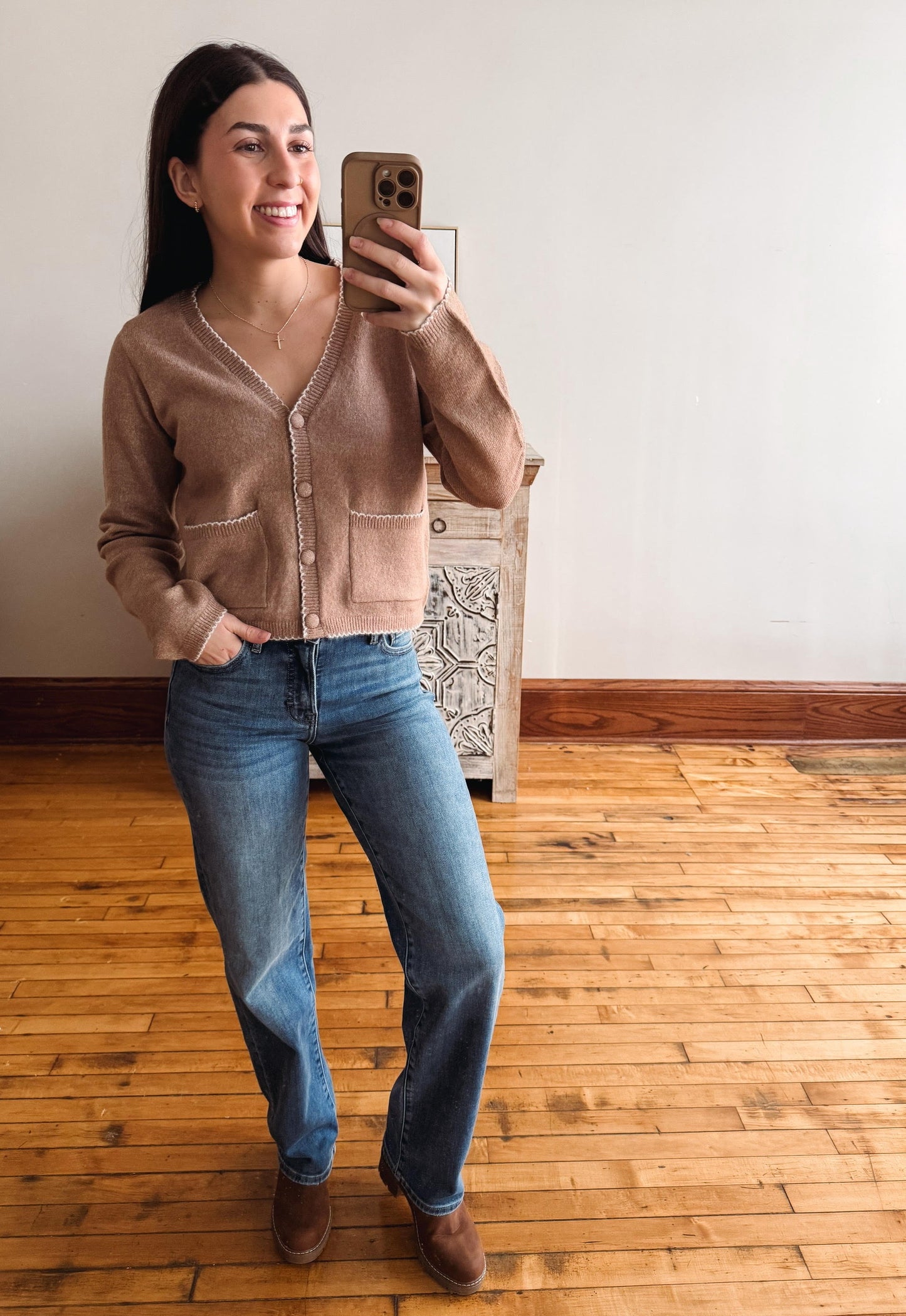 A woman takes a mirror selfie wearing a cropped brown knit cardigan, blue jeans, and brown boots in a room with a wooden floor and white wall.