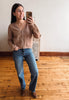 A woman takes a mirror selfie wearing a cropped brown knit cardigan, blue jeans, and brown boots in a room with a wooden floor and white wall.