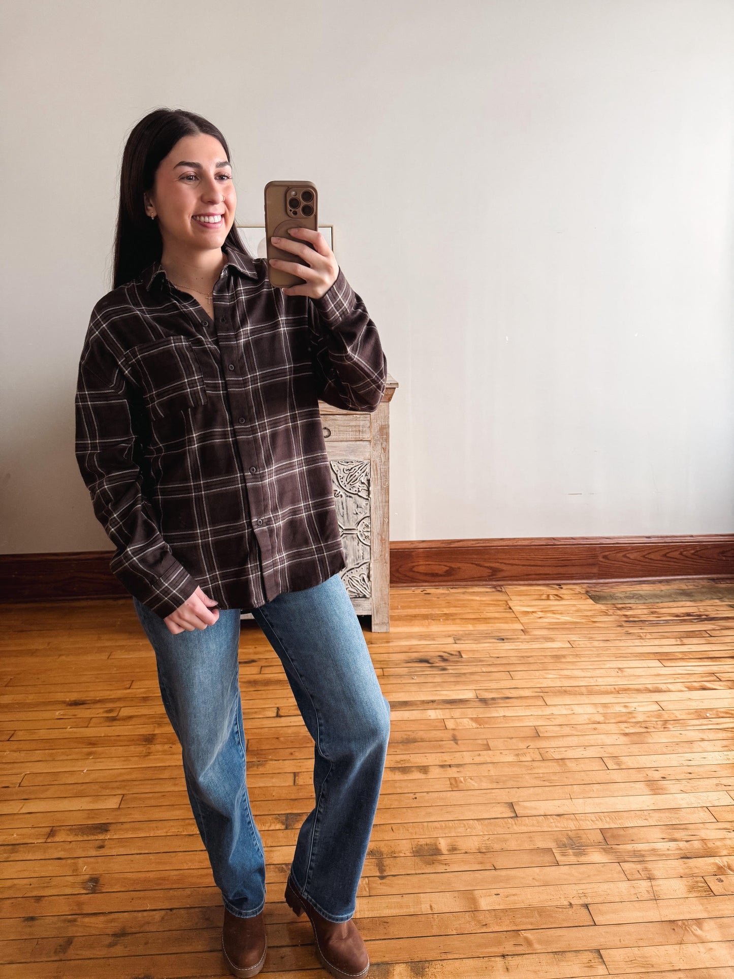 A woman taking a mirror selfie in a room with hardwood floors and a white wall, wearing a dark brown and black plaid flannel shirt, blue jeans, and brown boots.