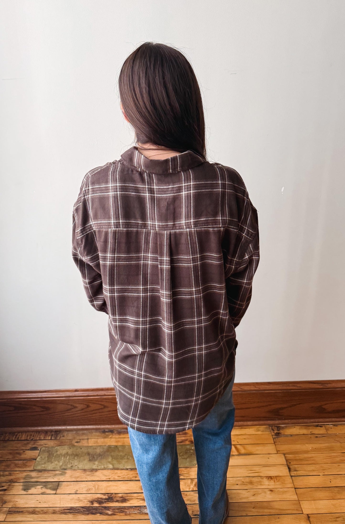A person viewed from behind wearing an oversized brown plaid button-up shirt and blue jeans, standing on a wooden floor against a plain off-white wall.