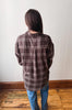 A person viewed from behind wearing an oversized brown plaid button-up shirt and blue jeans, standing on a wooden floor against a plain off-white wall.