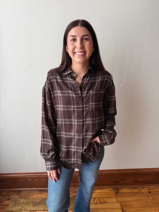 A woman with long dark hair stands indoors wearing a brown plaid button-up shirt and blue jeans, with a plain off-white wall and wooden floor behind her.